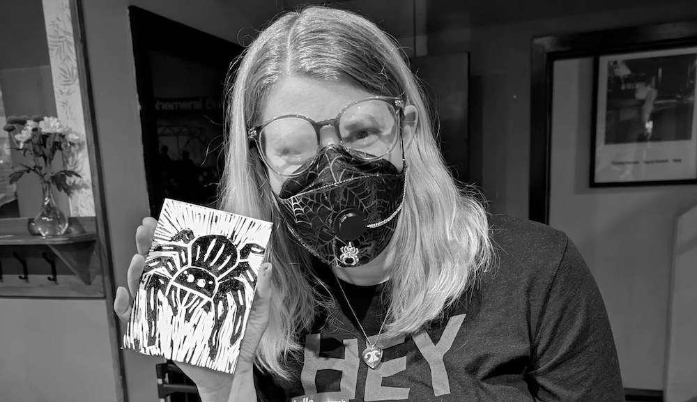 A person in a mask holding a lino cut of a spider