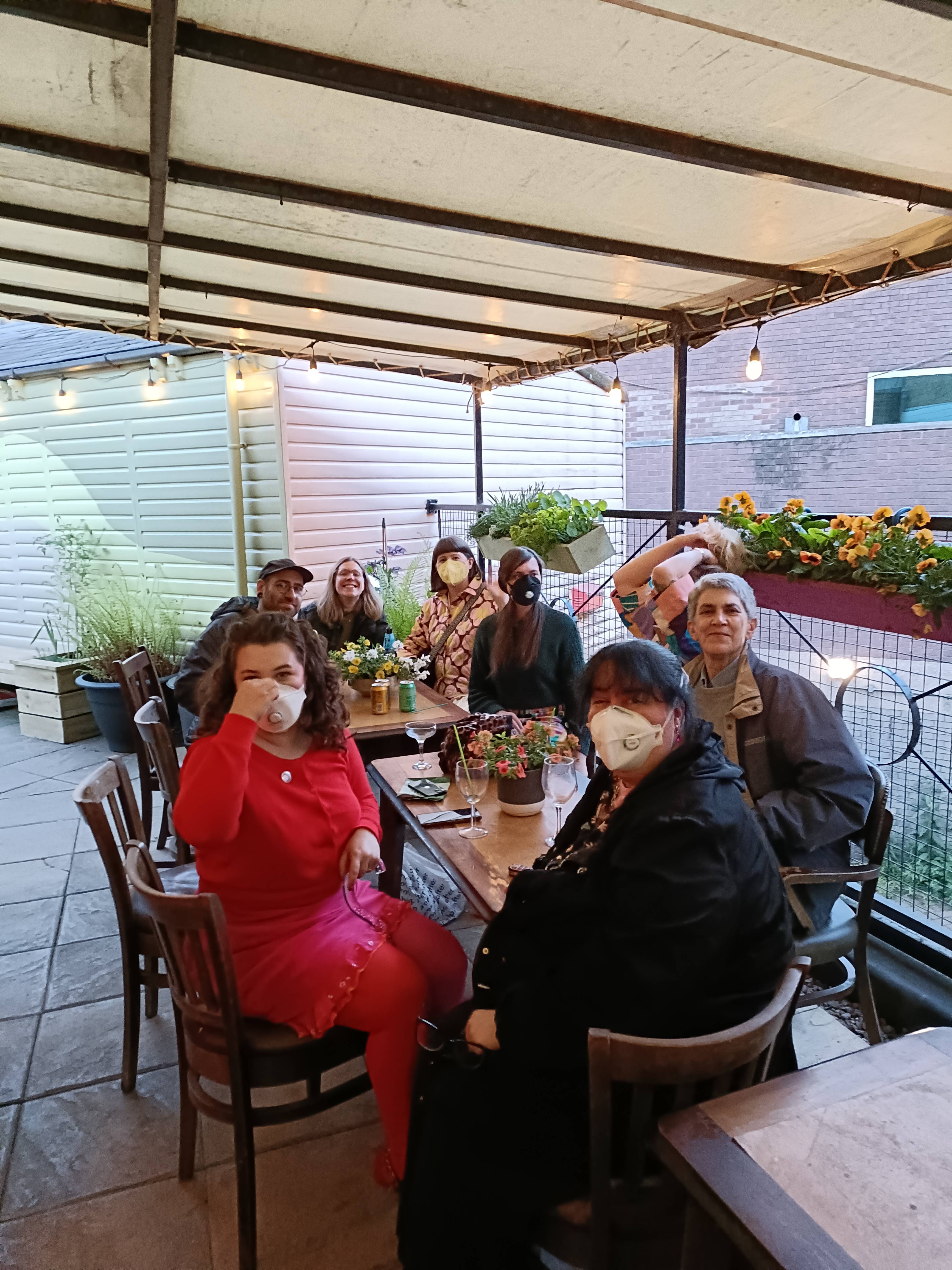 Summer social A group of people outside sitting around a table, most people are wearing masks and there are flowers in the background in nice pots
